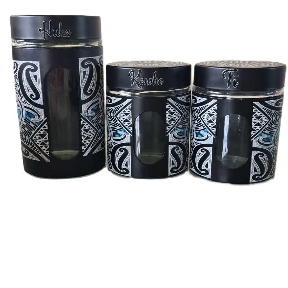 Three black canisters with Maori designs and glass windows. The lids are black with silver lettering reading 'Huka" 'Kawhe" "Ti" 