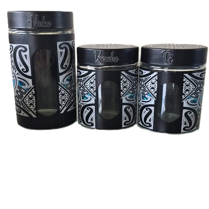 Three black canisters with Maori designs and glass windows. The lids are black with silver lettering reading 'Huka" 'Kawhe" "Ti" 